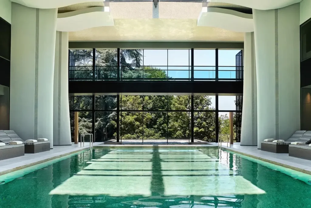 A green-tiled indoor pool faces tall windows overlooking trees, with pale loungers along the sides.