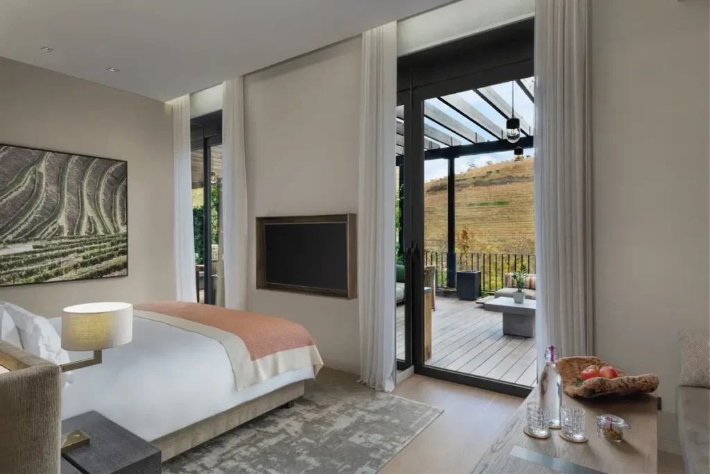 A pale bedroom with a vineyard artwork opens through glass doors onto a timber terrace facing vines.