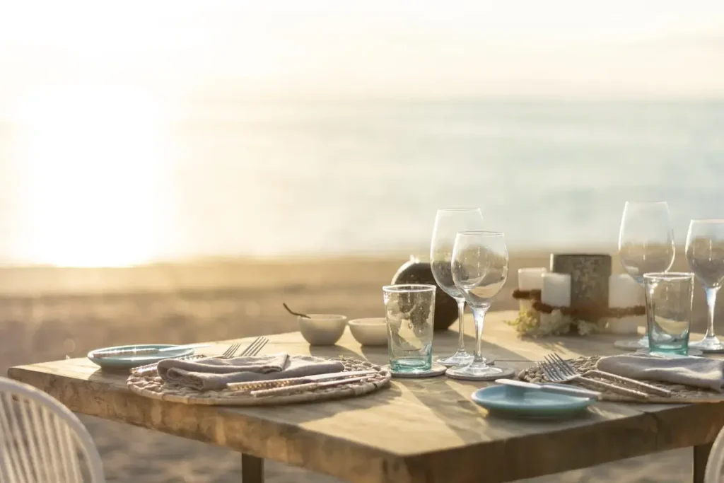 A wooden table set with plates, wine glasses, and candles faces softly lit sand and sea.