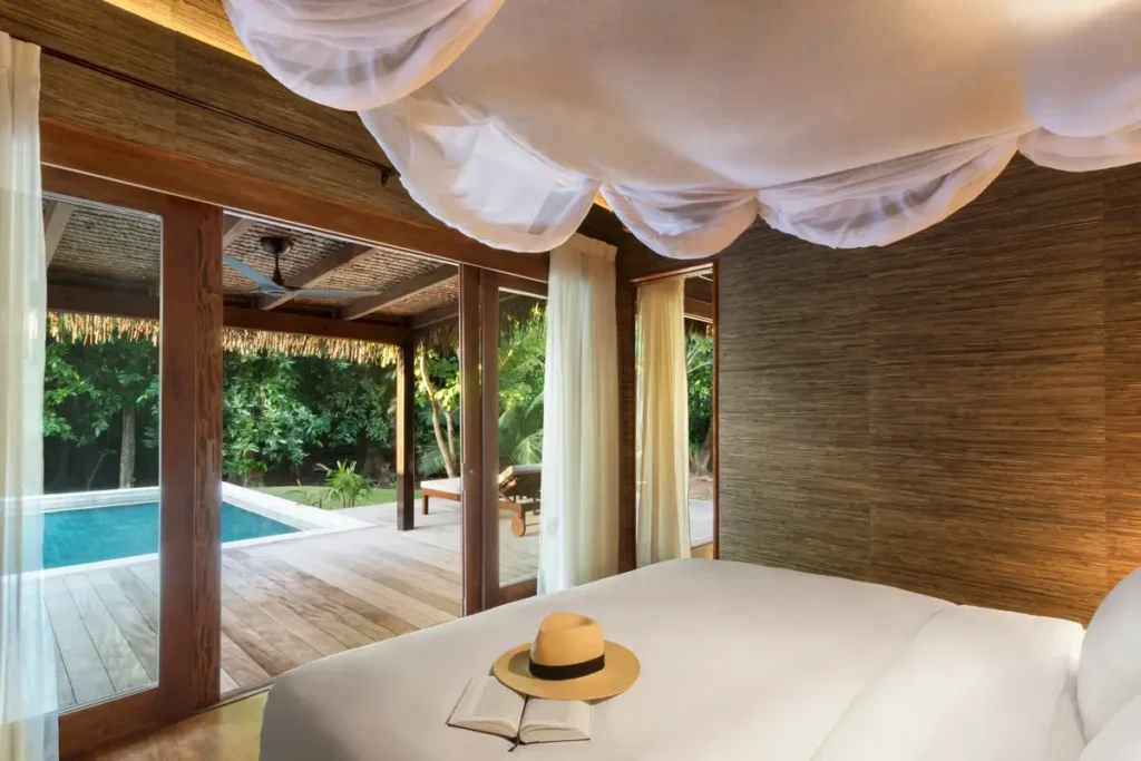 A bed beneath white draped fabric faces open timber-framed doors leading to a private pool and garden deck.