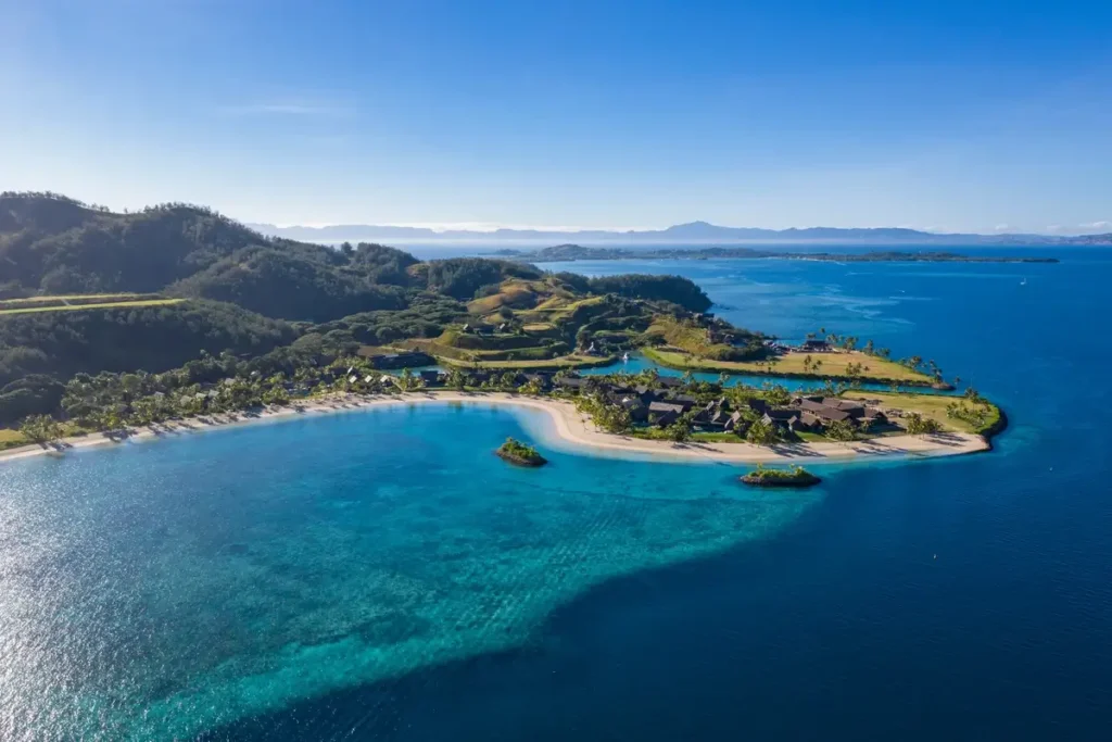 A turquoise bay borders a green coastal headland with resort buildings and hills rising behind it.