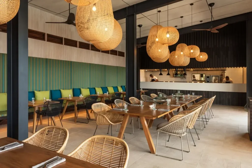 Woven pendant lights hang above wooden tables, rattan chairs, and a colorful banquette beside a restaurant bar.