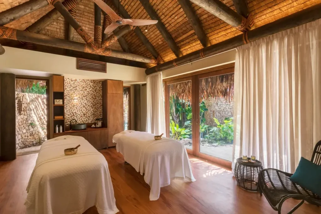 Two massage beds stand beneath a high timber roof beside curtains and windows facing tropical plants.