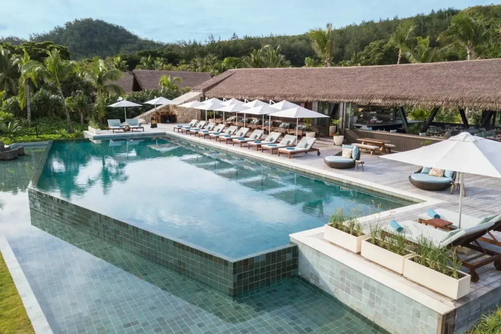 A broad swimming pool is bordered by loungers, white umbrellas, and thatched resort buildings beneath green hills.