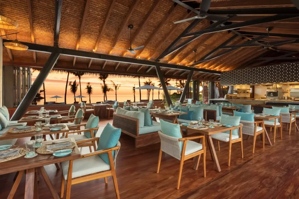 Set dining tables and pale blue chairs sit beneath an exposed timber roof facing palms and a sunset sky.