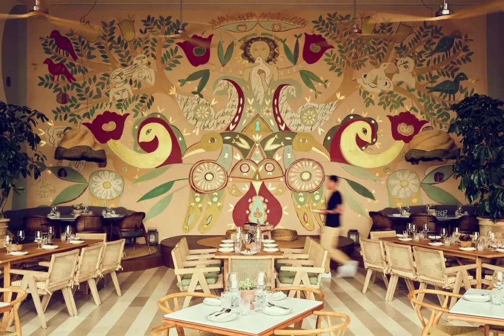 Set tables and timber chairs beneath a colorful mural in the Beach Caves dining room at Six Senses Ibiza
