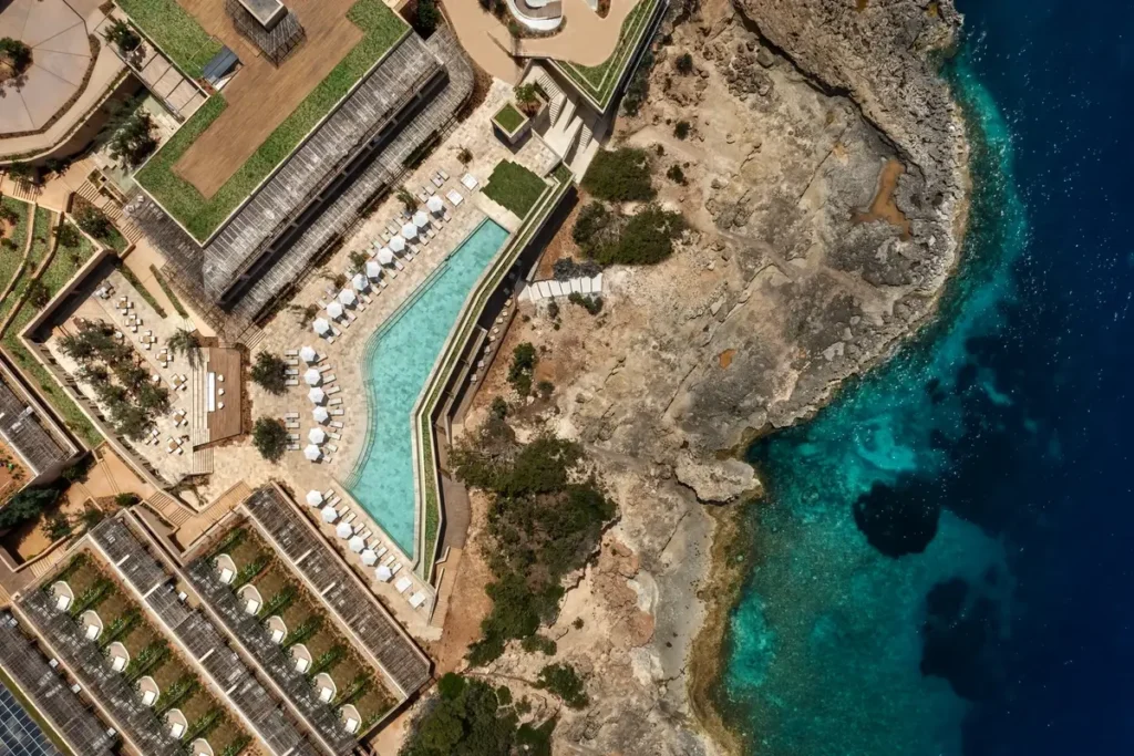 Overhead view of an angular pool, loungers, and rocky shoreline beside blue water at Six Senses Ibiza