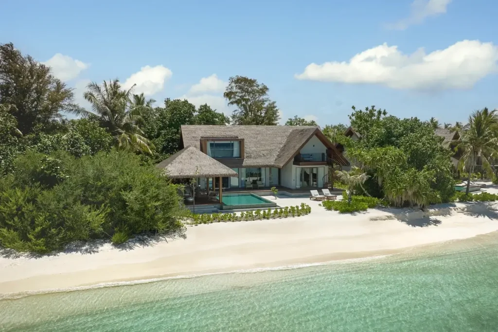 A beachfront retreat with a private pool stands among tropical trees behind white sand and clear lagoon water.