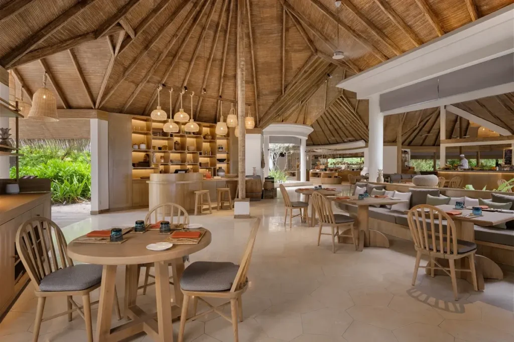 Wooden dining tables and chairs stand beneath a high exposed timber roof in a bright open restaurant.