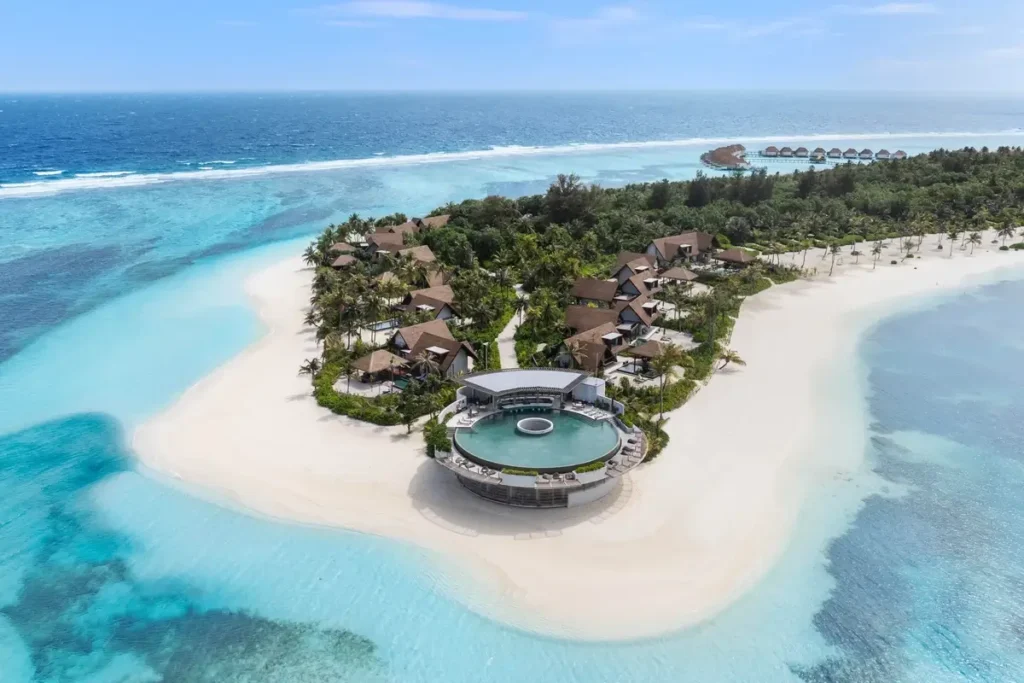 A raised circular pool sits at the edge of a broad beach, with villas, palms, and a turquoise lagoon behind it.