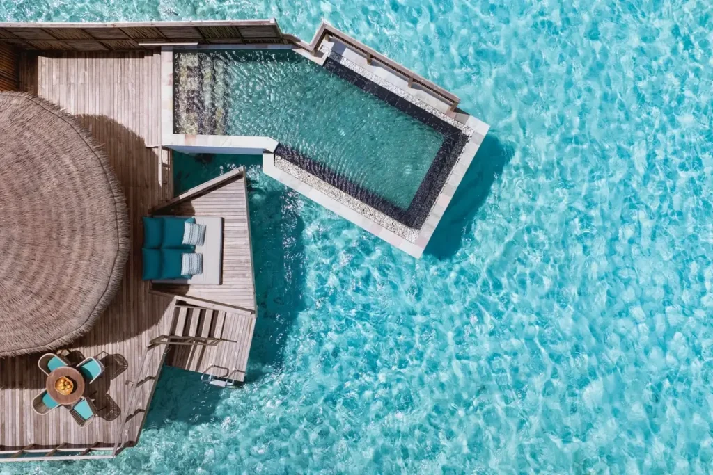 An overhead view shows a private pool projecting from a timber villa deck above bright turquoise water.