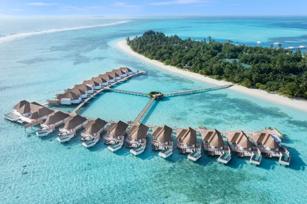 A curved row of thatched overwater villas extends into a turquoise lagoon beside a palm-covered island and white beach.