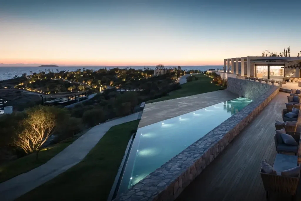 A long pool borders landscaped resort grounds, with hillside buildings and the sea beneath an evening sky.