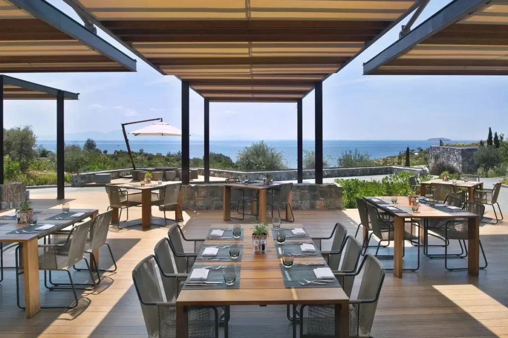 Set dining tables and woven chairs sit beneath timber shade structures on a terrace facing the sea.