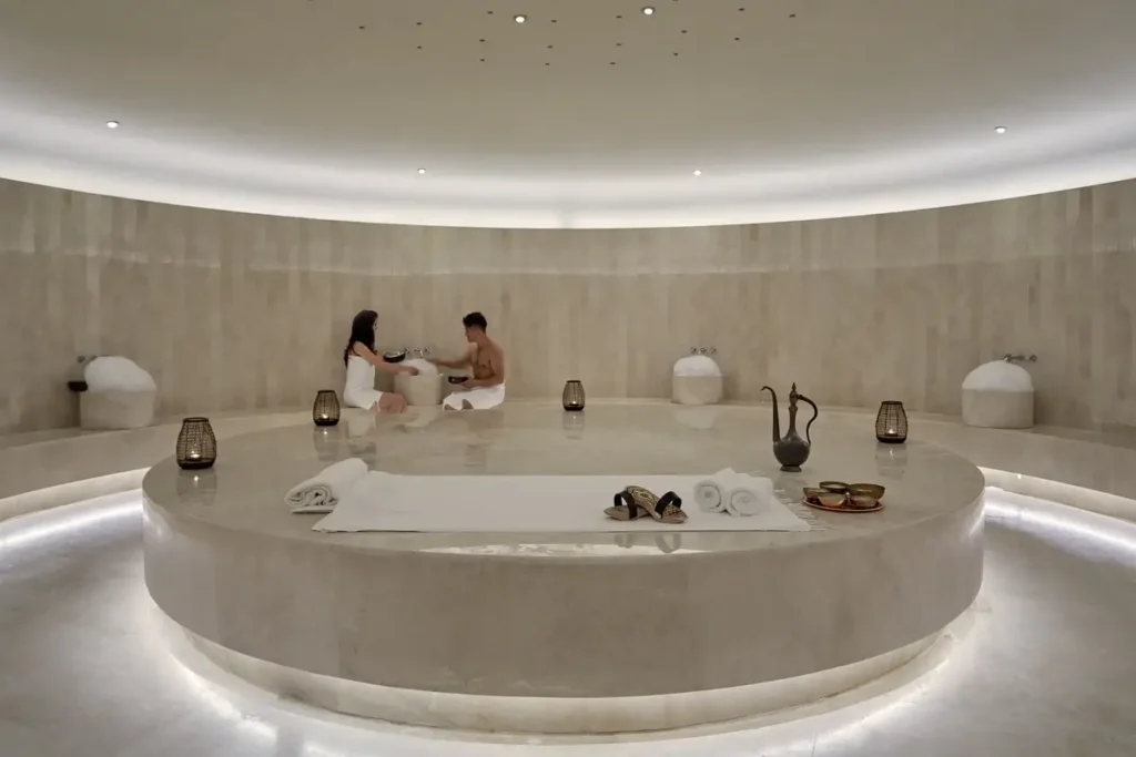 A circular pale-stone hammam has a central platform, bathing basins, and a treatment taking place at the far wall.