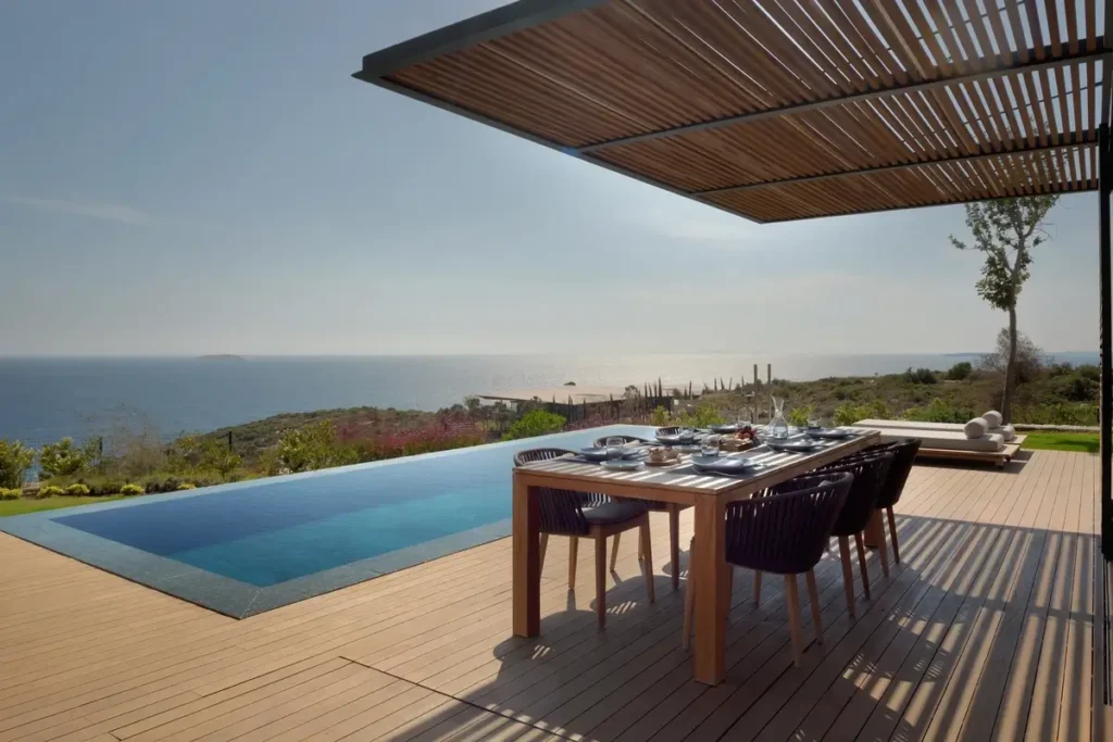 A set dining table stands beneath a slatted pergola beside a private pool overlooking the sea.