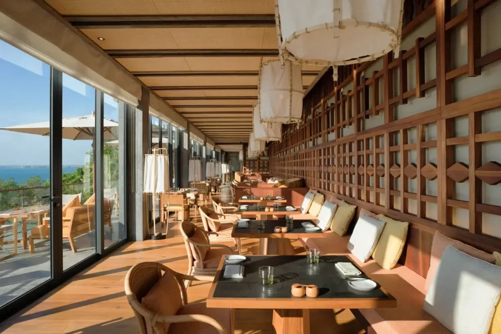 Rows of wooden tables and cushioned seating line a sunlit restaurant with large windows and pendant lights.