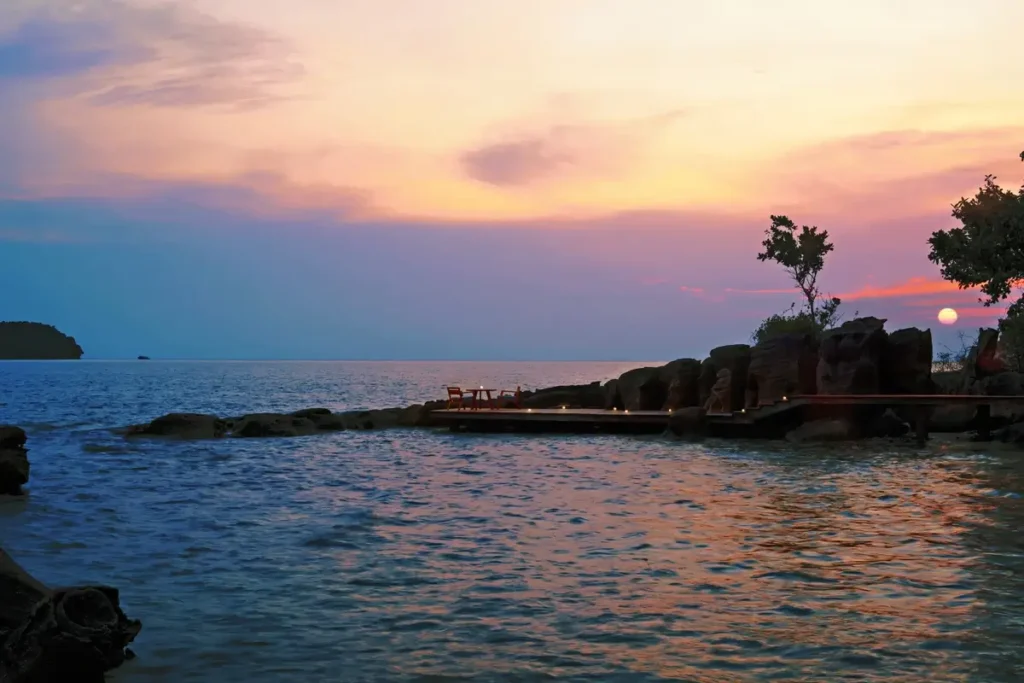 A low wooden boardwalk follows a rocky shoreline above the sea beneath a pink and orange evening sky.