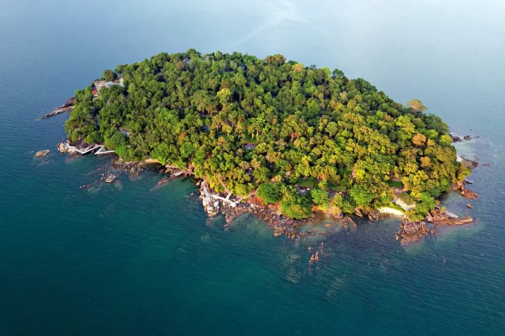 A small, densely forested island with a rocky shoreline is surrounded by blue water.