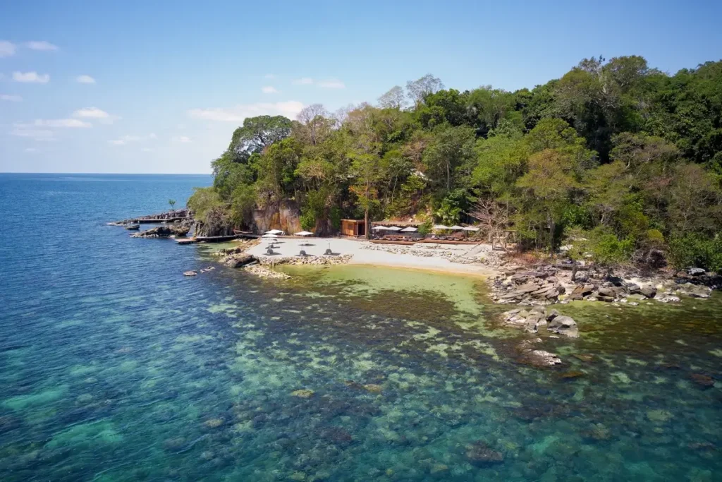 A small sandy beach with loungers curves between rocky shoreline and dense trees beside clear shallow water.