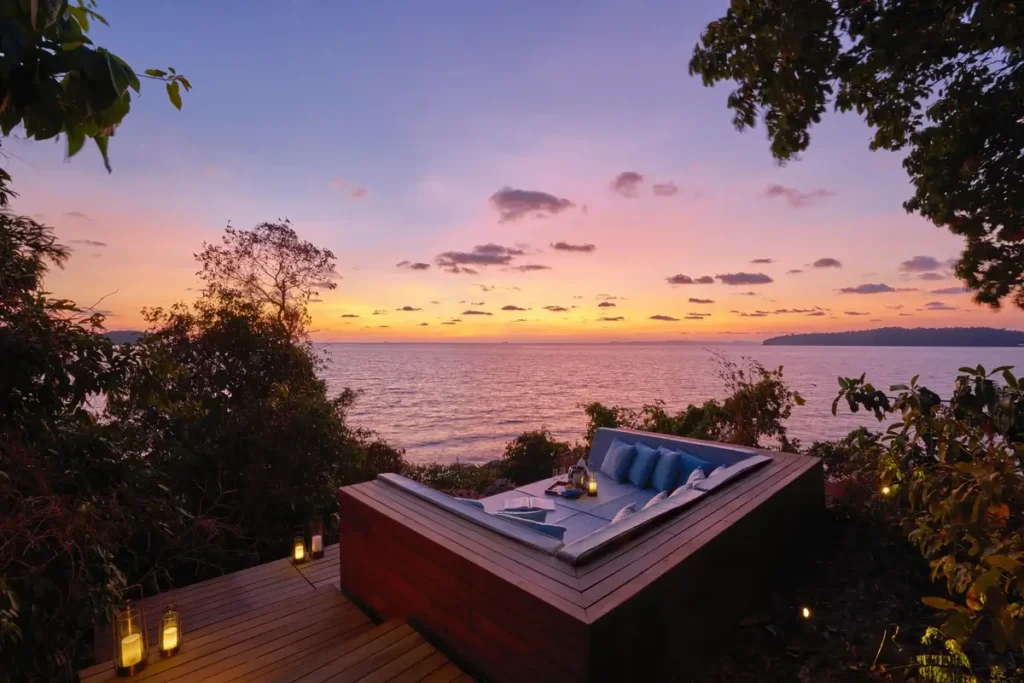 A cushioned daybed on a timber sundeck faces the sea beneath a pink and orange sunset sky.