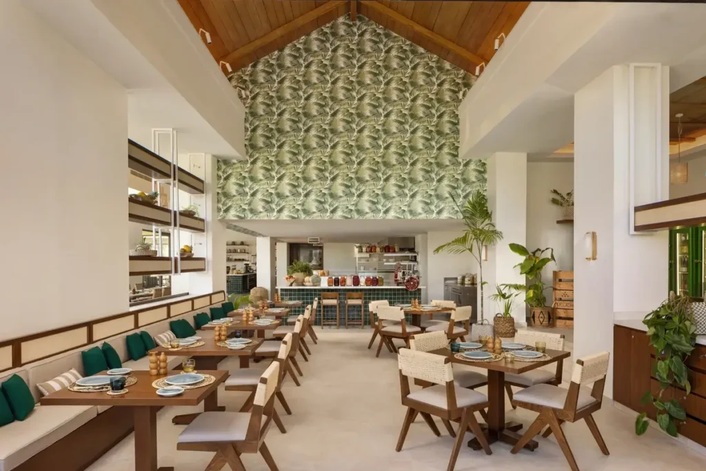 Wooden dining tables and green-cushioned seating fill a bright restaurant beneath a high timber roof and patterned wall.