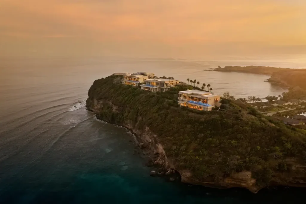 Villas sit on a green coastal headland above blue water as a warm sunset lights the surrounding bay.