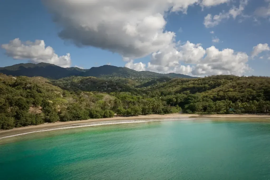 Turquoise water curves around a sandy bay backed by dense greenery and distant hills beneath a cloudy blue sky.