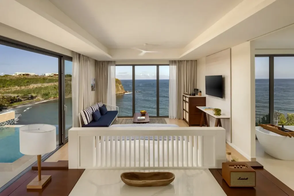 A bright suite with a white bed, blue sofa, and large windows looks toward the sea and a green headland.