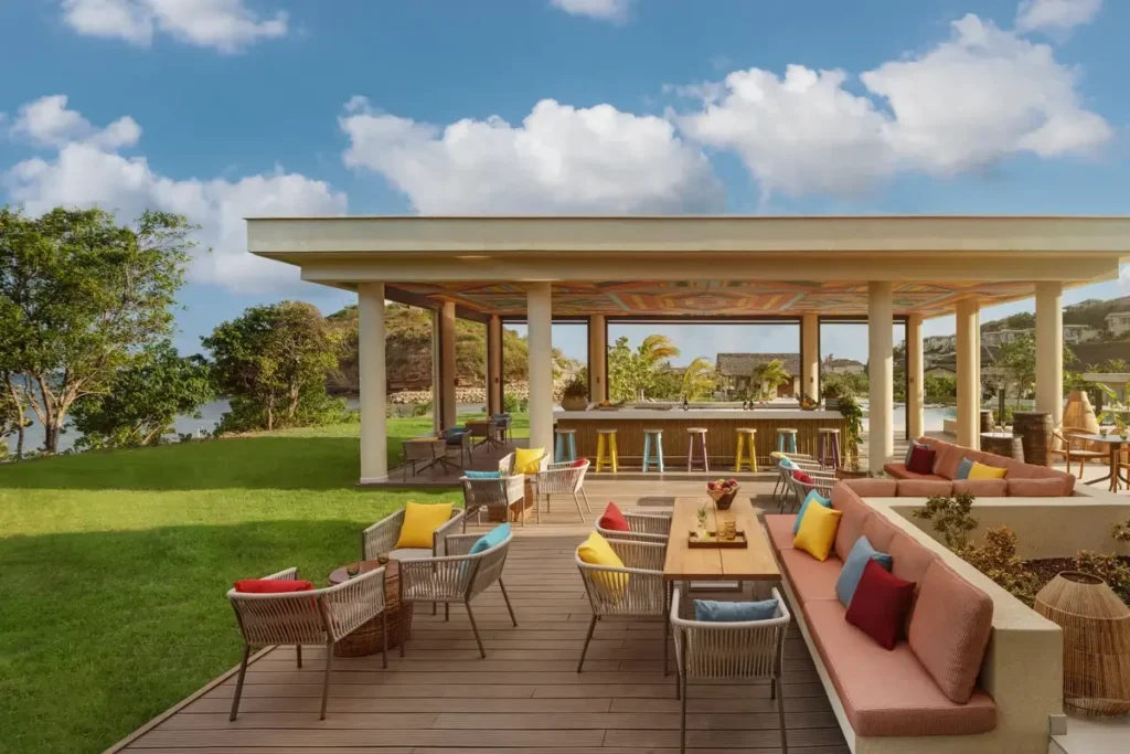 Colorful chairs and cushioned benches surround outdoor tables beside a green lawn and an open pavilion bar.