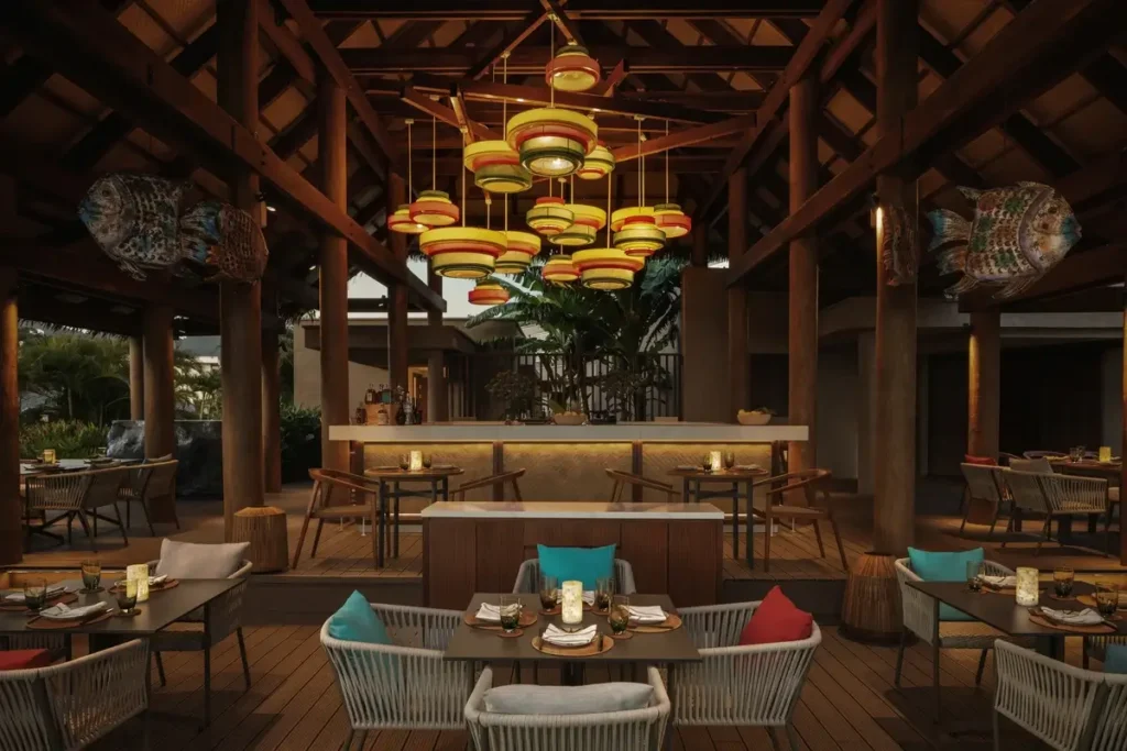 Colorful woven pendant lights hang above a restaurant bar and dining tables beneath a dark timber roof.