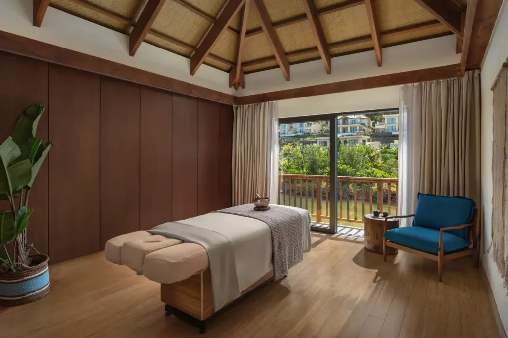 A massage bed stands beneath an exposed timber roof beside a blue chair and glass doors opening to a balcony.