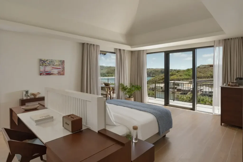 A pale bedroom with a blue bed runner and wooden desk opens toward a balcony overlooking greenery and resort buildings.