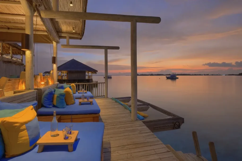Blue-cushioned seats and low tables line a wooden overwater deck facing calm water and an orange sunset.