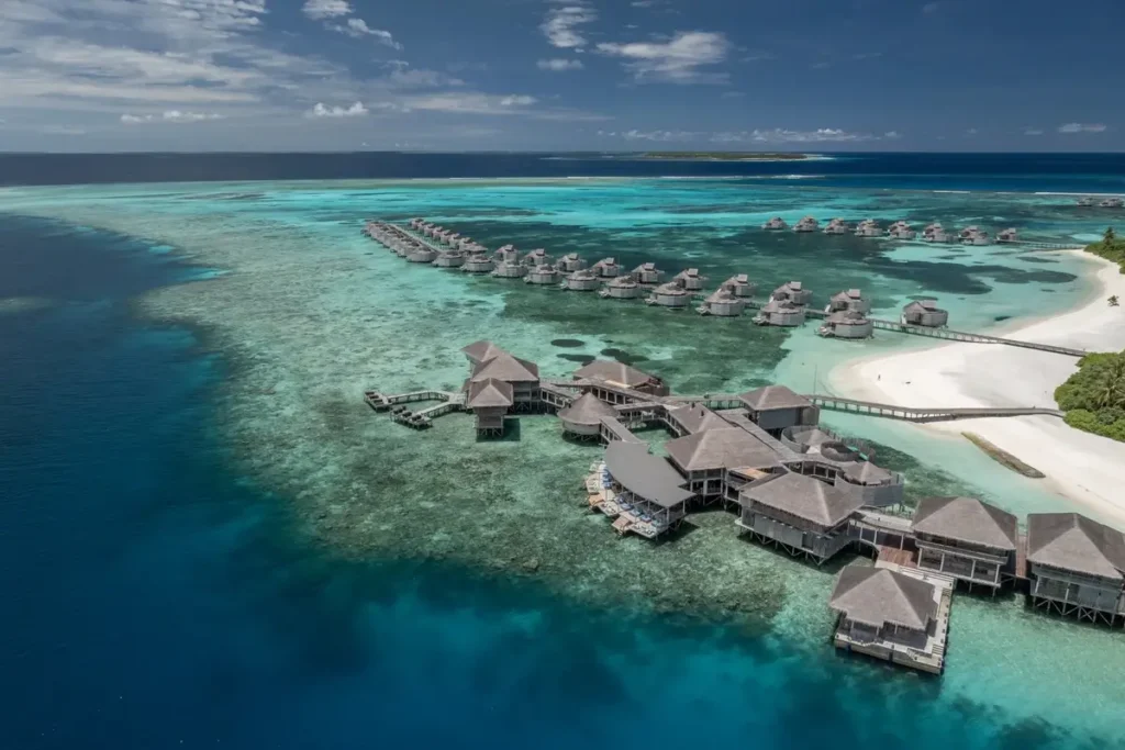 Thatched water villas follow wooden jetties across turquoise water beside a white-sand shoreline at Six Senses Laamu.