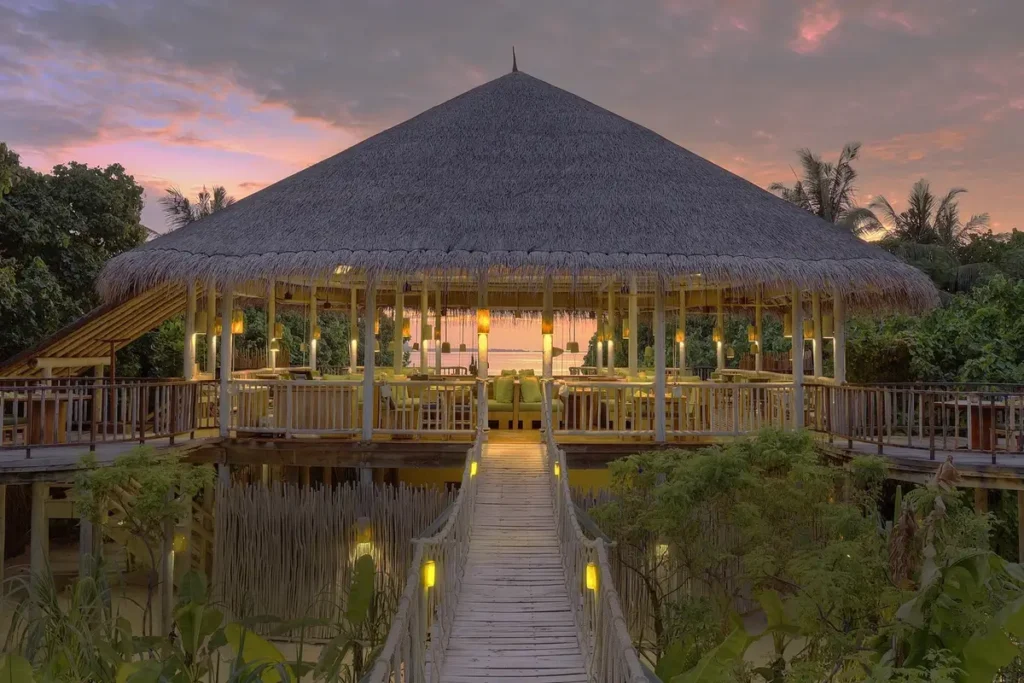 A wooden walkway leads to the illuminated thatched pavilion of Leaf restaurant beneath a pink evening sky.
