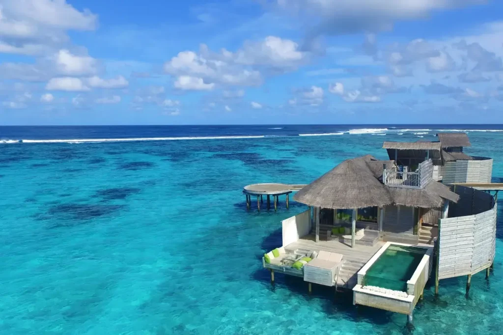 A thatched overwater villa has a private pool and wooden deck above turquoise water at Six Senses Laamu.