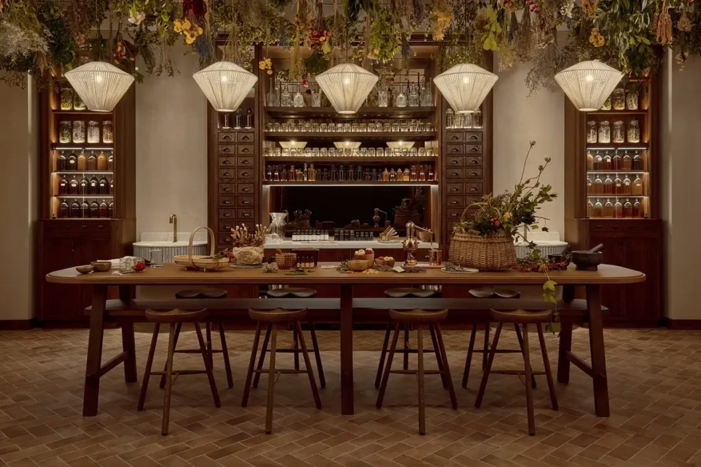 A long wooden table and chairs sit before shelves of jars beneath pendant lights and hanging dried plants.