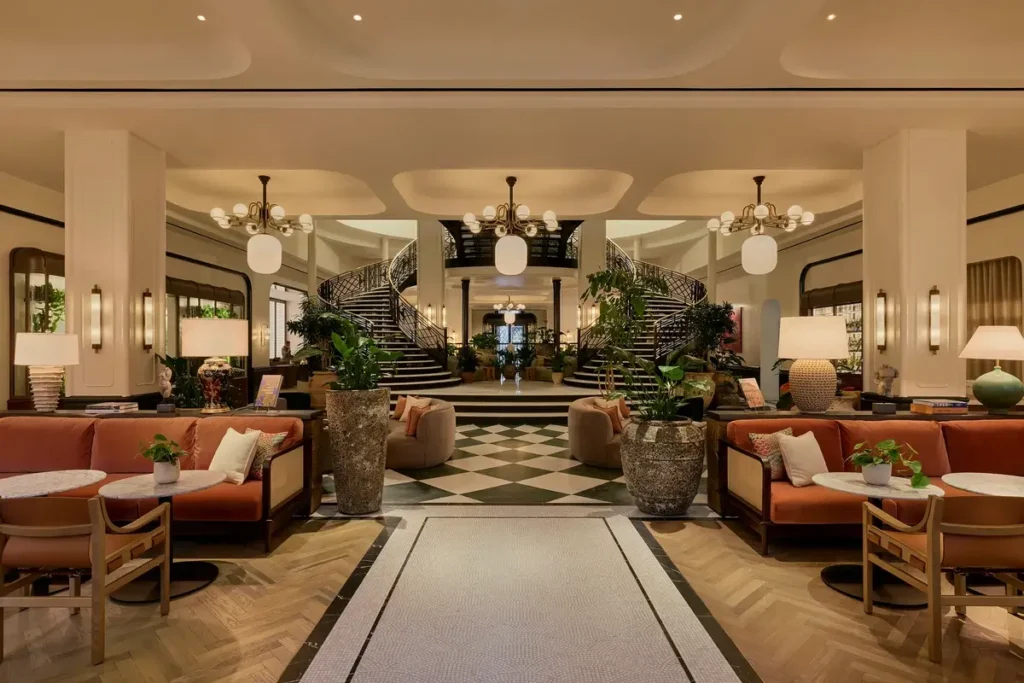 Curved twin staircases rise beyond patterned floors, large plants, and cushioned lounge seating in the hotel courtyard.