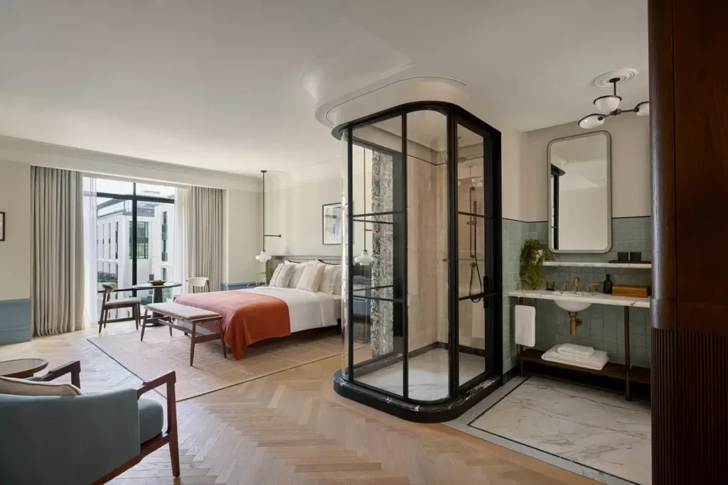 A glass-enclosed shower and adjacent vanity stand beside the sleeping area in a light-filled Premier room.