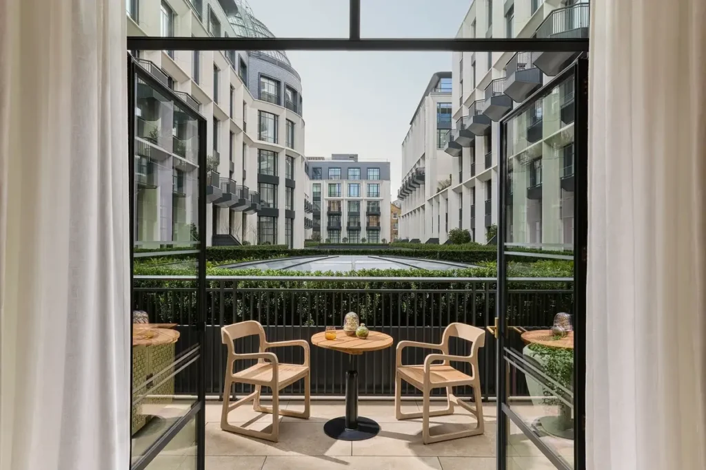 Two timber chairs and a round table sit on a private terrace facing a landscaped courtyard between buildings.