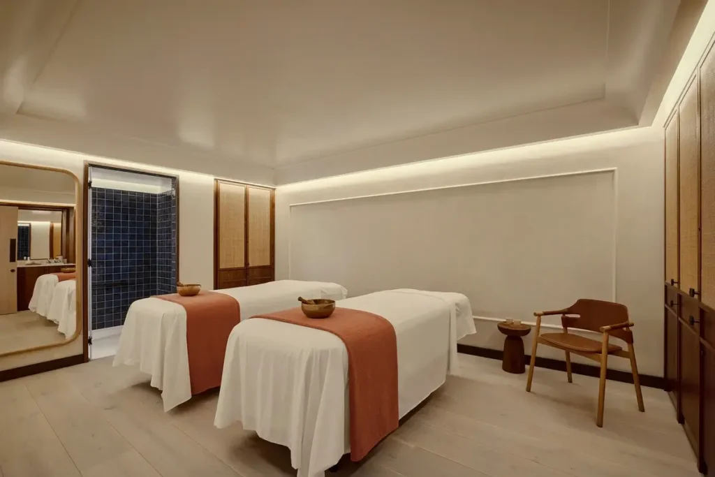 Two white-covered treatment tables with rust-colored runners stand in a softly lit room with a timber chair.