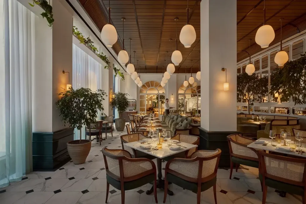 Set dining tables, upholstered chairs, and tall plants fill a restaurant with globe pendant lights and large windows.