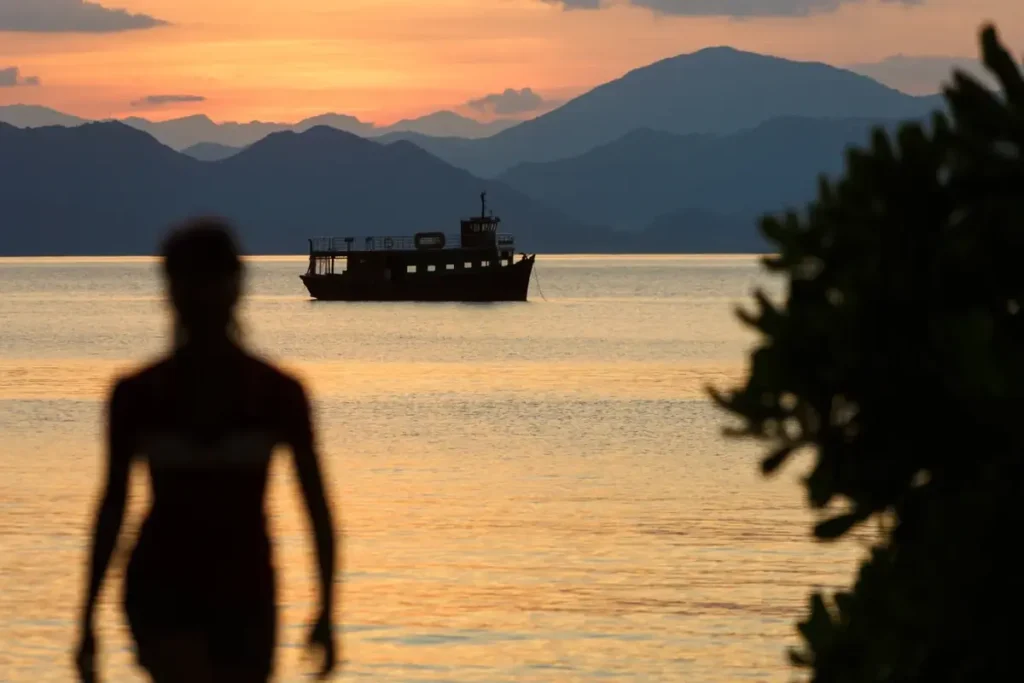 Silhouetted person facing a boat on golden water beneath mountain ridges at Six Senses Ninh Van Bay