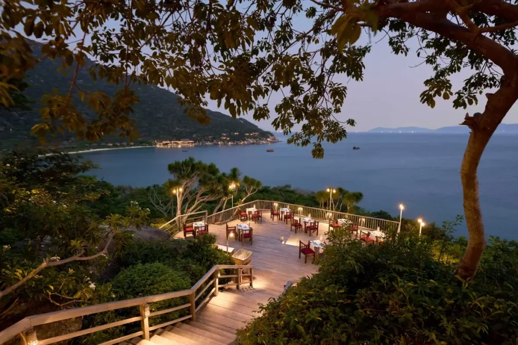Outdoor dining terrace with set tables above a wooded shoreline and blue bay at Six Senses Ninh Van Bay