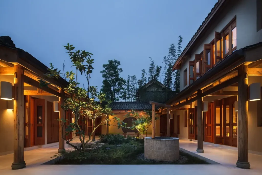 Warmly lit two-story buildings surround a planted courtyard with a circular stone feature beneath a blue evening sky.