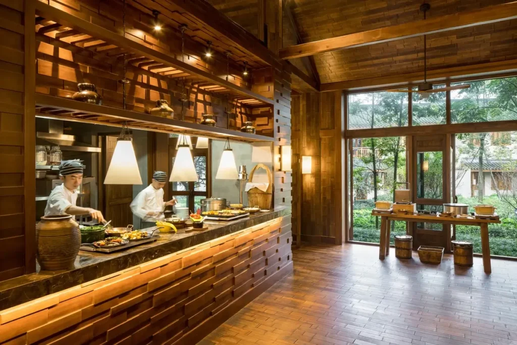 Chefs work behind a long wooden counter beneath pendant lights and exposed beams beside a bright garden-facing dining space.