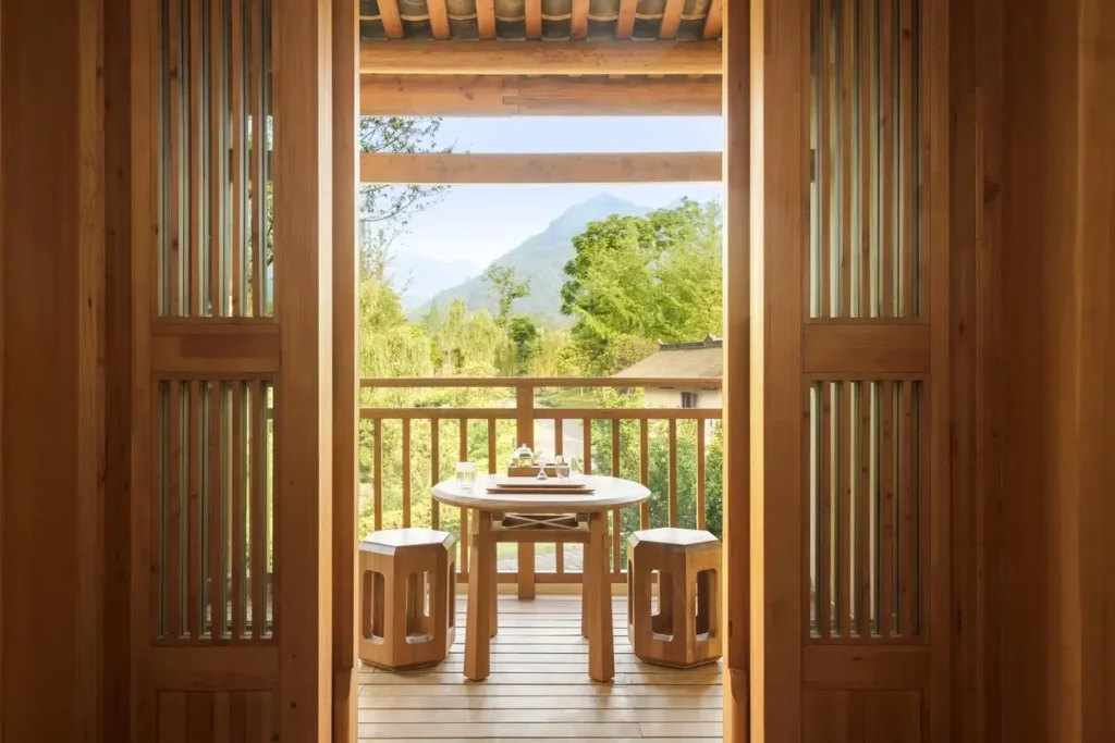 Open wooden doors frame a balcony with a round table and stools facing trees and a distant mountain.