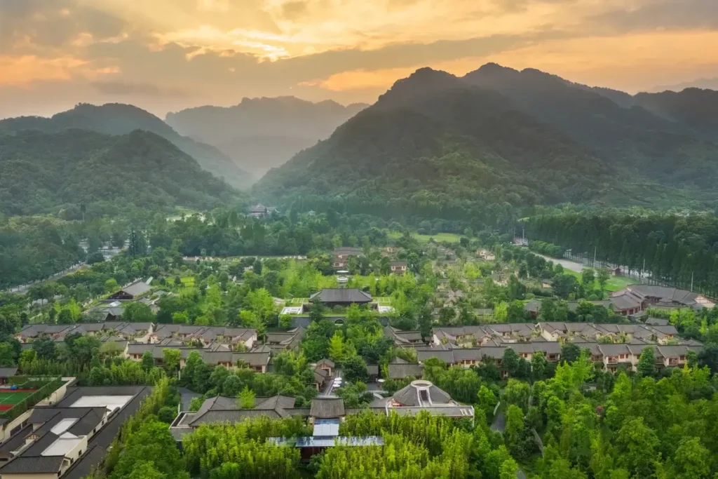 Low-rise resort buildings and green courtyards spread beneath forested mountains under a golden evening sky.
