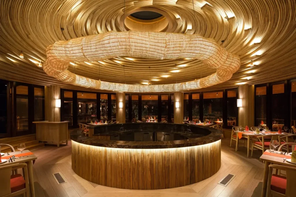 A circular counter sits beneath a large woven light fixture and curved timber ceiling in a warmly lit restaurant.